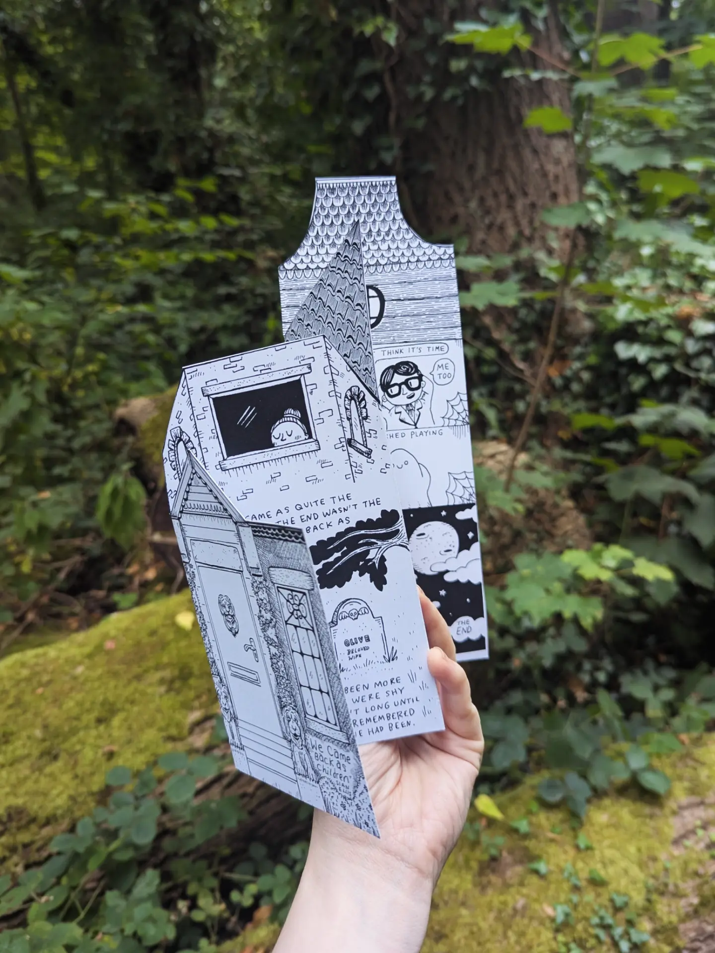 A hand holding a hand-folded haunted house zine in the woods